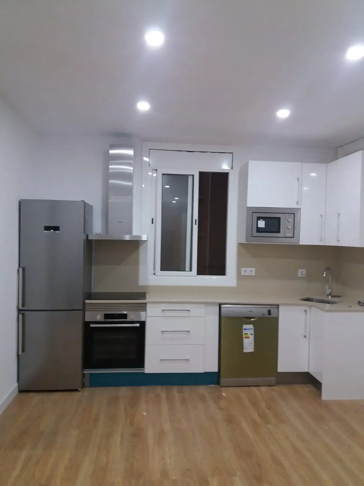 Cocina abierta blanca con electrodomésticos de acero inoxidable, suelo de madera y focos empotrados