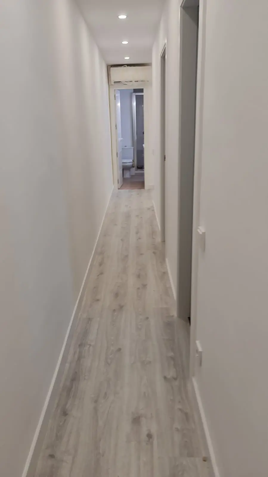 Pasillo con suelo laminado gris, paredes pintadas en blanco y focos empotrados